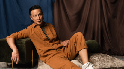 Daniel Wu posted in a tan jumpsuit in front a multi-colored curtain.