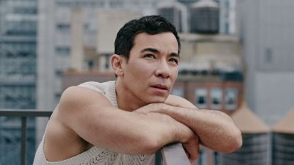 Conrad Ricamora leaning on a balcony while staring at the New York City Skyline