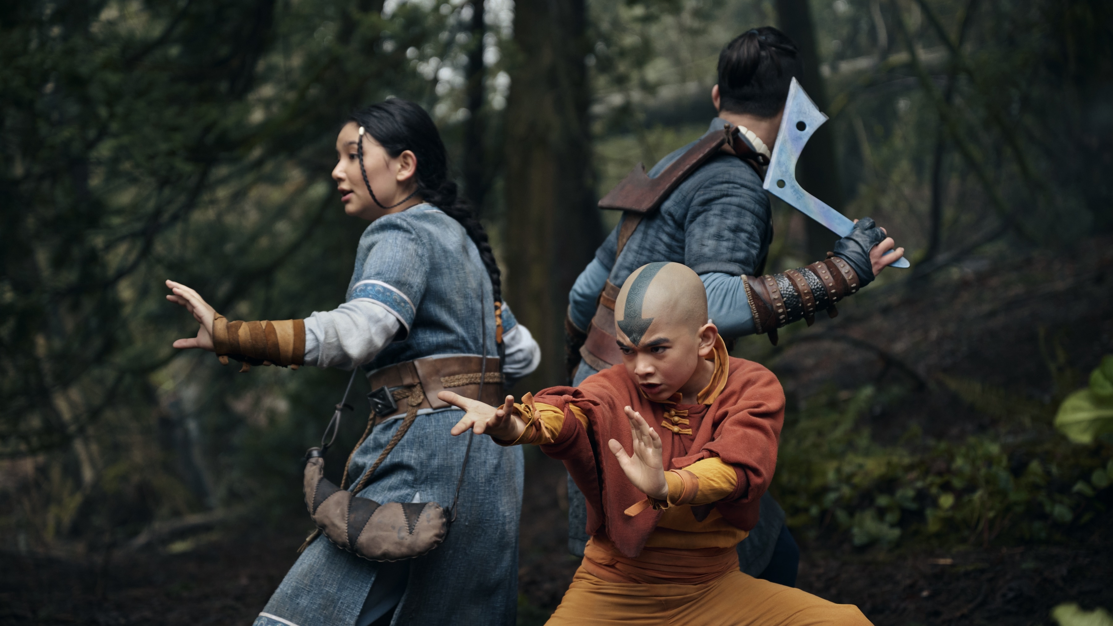 Katara (Kiawentiio), Aang (Gordon Cormier) and Sokka (Ian Ousley) stand back to back in a forest.
