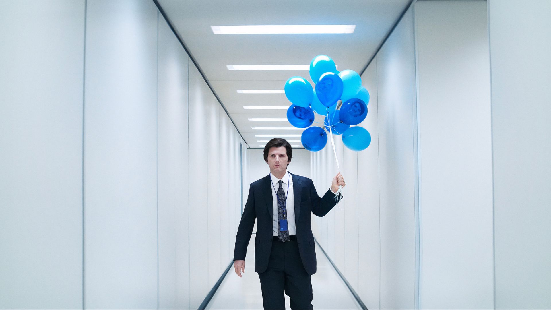 Mark Scout (Adam Scott) walks down an all-white hallway wearing a suit and carrying blue balloons.